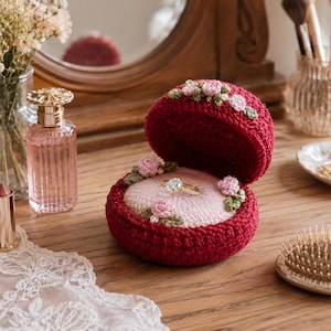 May include: A red crocheted ring box, open to display a diamond ring on a pink cushion. The box is decorated with small pink flowers and green leaves. Other items on the wooden surface include perfume, lipstick, and a hairbrush.