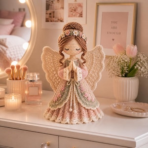 May include: A handcrafted crochet angel doll with a halo of flowers, closed eyes, and folded hands. The doll wears a pink, green, and cream-colored dress with lace wings. The angel is displayed on a white surface with a mirror and other decorative items.