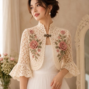 May include: A cream-coloured crochet bolero jacket featuring a high collar, three-quarter length bell sleeves, and a decorative metal clasp closure. The garment is adorned with intricate pink and green floral embroidery on both front panels.