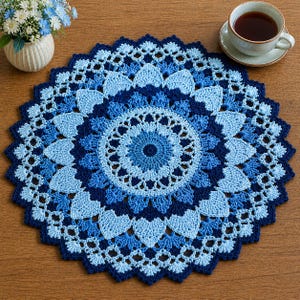 May include: A round, blue crochet doily with a floral design. The doily features concentric circles of varying shades of blue, from light to dark. The edges have a scalloped trim. A cup of tea and a small vase of flowers are in the background.