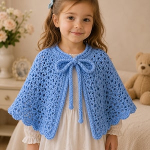 Può includere: Una mantella all'uncinetto blu chiaro, con apertura frontale, bordo smerlato e chiusura a fiocco. La mantella fatta a mano è indossata sopra un vestito bianco. La mantella ha un design delicato e arioso, perfetto per un bambino.