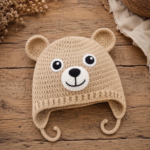 May include: A tan crocheted bear hat with a white and black bear face. The hat has ear flaps and small, rounded ears. The hat is made of a soft, textured yarn and is designed for babies or young children.