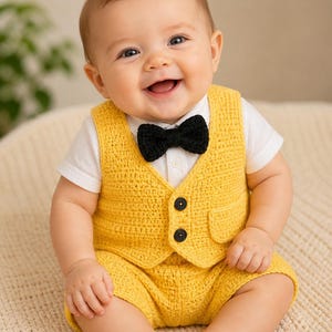 Puede incluir: Un bebé con un conjunto de chaleco y pantalón corto de crochet amarillo, con una pajarita negra. El conjunto incluye una camisa blanca de manga corta. El bebé sonríe y está sentado sobre una superficie color crema.