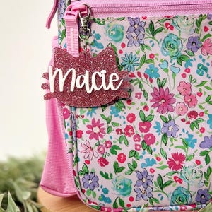 May include: Pink glitter cat-shaped name tag with the name "Macie" written in white. The tag is attached to a pink floral backpack.