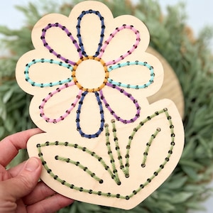 K&ouml;nnte beinhalten: Holzblumen-Bastelset mit vorgebohrten L&ouml;chern zum Einf&auml;deln von Garn. Die Blume hat eine violette, rosa, blaue und gr&uuml;ne Mitte mit gr&uuml;nen Bl&auml;ttern.