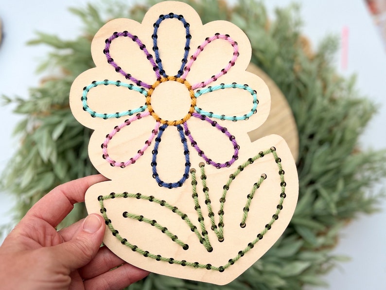 K&ouml;nnte beinhalten: Holzblumen-Bastelset mit vorgebohrten L&ouml;chern zum Einf&auml;deln von Garn. Die Blume hat eine violette, rosa, blaue und gr&uuml;ne Mitte mit gr&uuml;nen Bl&auml;ttern.
