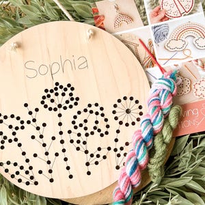 May include: A wooden embroidery kit with a floral design and the name "Sophia" written on it. The kit includes two skeins of yarn, one pink and white and one green.