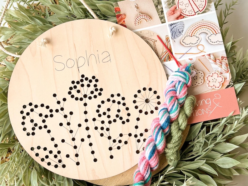 May include: A wooden embroidery kit with a floral design and the name "Sophia" written on it. The kit includes two skeins of yarn, one pink and white and one green.