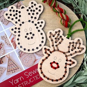 May include: Wooden reindeer ornaments with pre-drilled holes for yarn sewing. One reindeer is partially sewn with brown yarn, the other is ready to be sewn. A booklet with instructions and other Christmas-themed sewing projects is also visible.