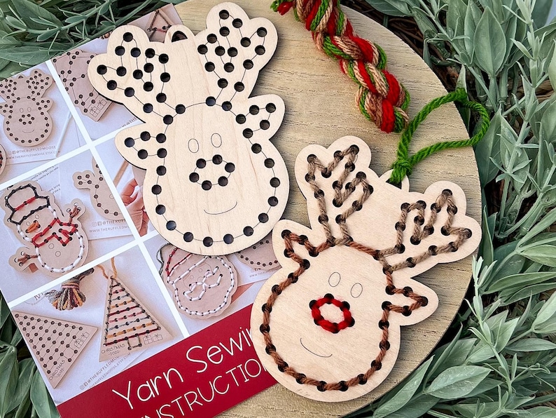 May include: Wooden reindeer ornaments with pre-drilled holes for yarn sewing. One reindeer is partially sewn with brown yarn, the other is ready to be sewn. A booklet with instructions and other Christmas-themed sewing projects is also visible.