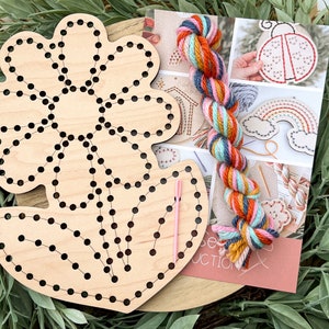 K&ouml;nnte beinhalten: Ein Holz-Stickset mit einer vorgefertigten Blumenform, einer Nadel und buntem Garn. Das Garn ist eine Mischung aus Orange, Rosa, Blau und Gelb. Das Set enth&auml;lt Anleitungen zum Sticken einer Blume.