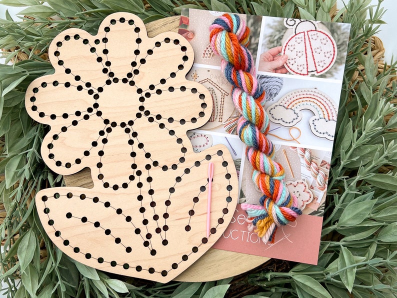 K&ouml;nnte beinhalten: Ein Holz-Stickset mit einer vorgefertigten Blumenform, einer Nadel und buntem Garn. Das Garn ist eine Mischung aus Orange, Rosa, Blau und Gelb. Das Set enth&auml;lt Anleitungen zum Sticken einer Blume.