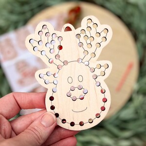 May include: Wooden reindeer Christmas ornament with a series of small holes filled with red, white, and brown beads. The reindeer has a smiling face and a loop for hanging. The ornament is on a wooden surface.