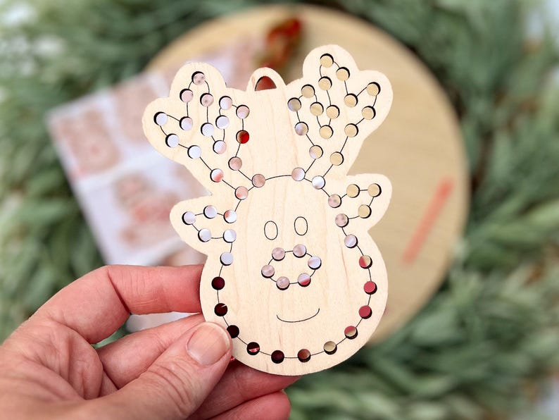 May include: Wooden reindeer Christmas ornament with a series of small holes filled with red, white, and brown beads. The reindeer has a smiling face and a loop for hanging. The ornament is on a wooden surface.