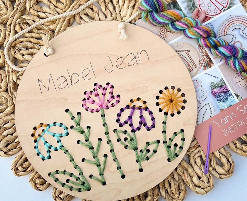 May include: A wooden embroidery hoop with the name "Mabel Jean" written on it. The hoop has a design of flowers and leaves that are ready to be embroidered with yarn.