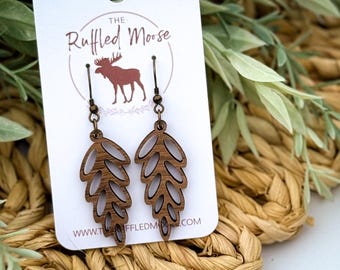 May include: Wooden leaf-shaped earrings with a cutout design. The earrings are a dark brown color and hang from bronze-colored ear wires. The earrings are displayed on a white card with the logo of "The Ruffled Moose".