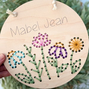 May include: A round wooden sign with the name "Mabel Jean" embroidered in black thread. The sign is decorated with four embroidered flowers in pink, purple, blue, and yellow.