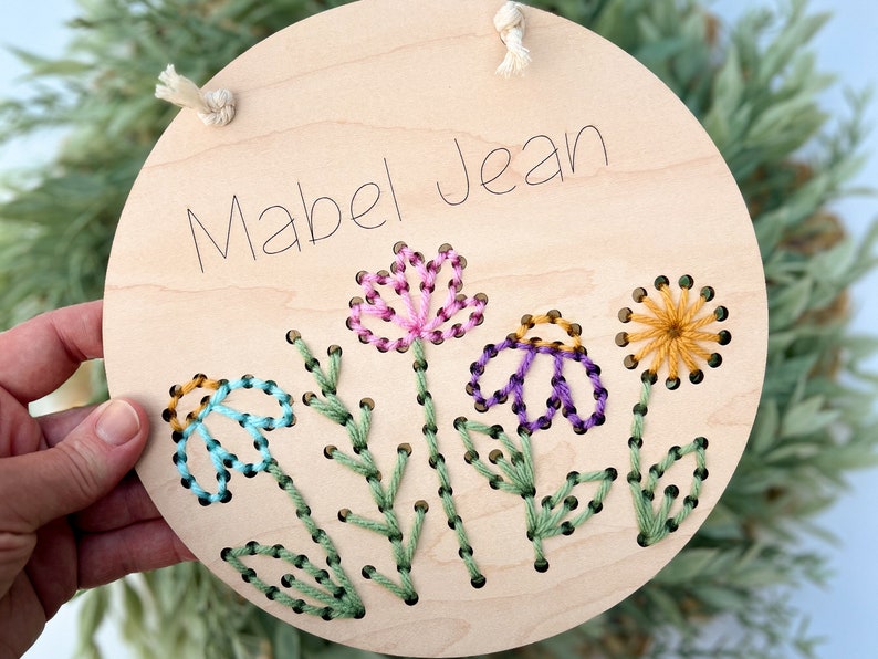 May include: A round wooden sign with the name "Mabel Jean" embroidered in black thread. The sign is decorated with four embroidered flowers in pink, purple, blue, and yellow.