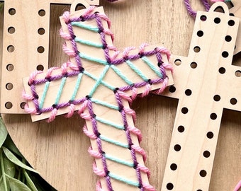 May include: A wooden cross with holes for stringing yarn. The cross is decorated with pink and teal yarn in a diagonal pattern.