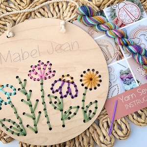 May include: A wooden embroidery hoop with the name "Mabel Jean" written on it. The hoop is decorated with a colorful flower design that has been stitched with yarn. The yarn and needle are included in the kit.