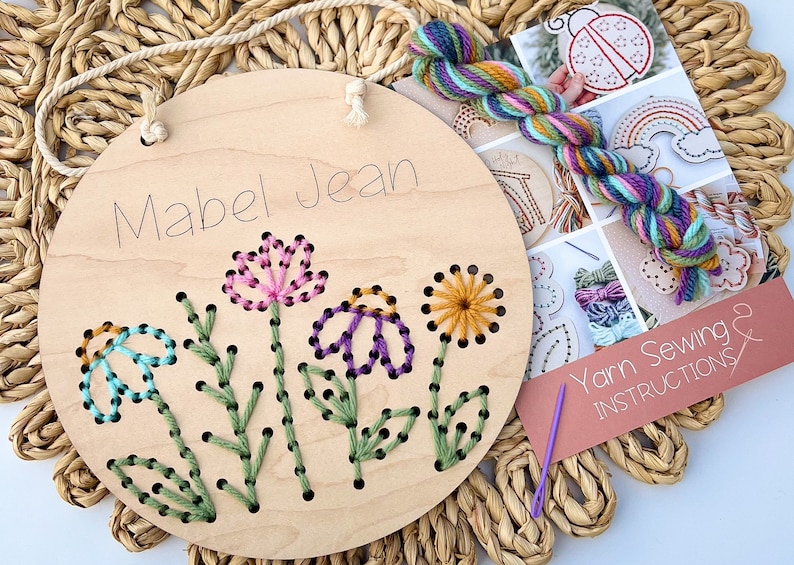 May include: A wooden embroidery hoop with the name "Mabel Jean" written on it. The hoop is decorated with a colorful flower design that has been stitched with yarn. The yarn and needle are included in the kit.
