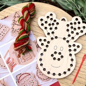 May include: A wooden reindeer-shaped craft with pre-drilled holes for string art, accompanied by a red needle and a skein of red, green, and brown yarn. Additional wooden ornaments and instructions are visible.