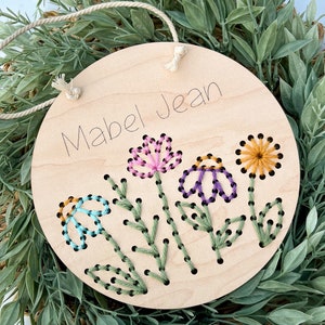 May include: A round wooden sign with the name "Mabel Jean" written on it. The sign is decorated with embroidered flowers in shades of pink, purple, blue, yellow, and green.