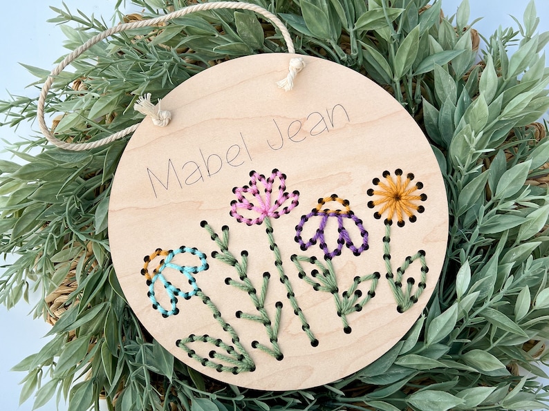 May include: A round wooden sign with the name "Mabel Jean" written on it. The sign is decorated with embroidered flowers in shades of pink, purple, blue, yellow, and green.