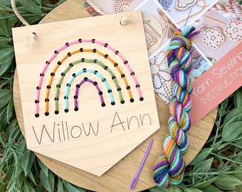 May include: A wooden rainbow craft kit with colorful yarn for stitching. The kit includes a wooden plaque with pre-punched holes for stitching a rainbow design. The plaque also has the name "Willow Ann" written on it.
