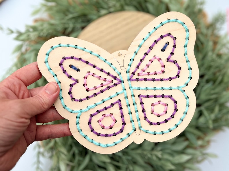 K&ouml;nnte beinhalten: Ein Holzschmetterling mit einer hellbraunen Beize, der mit einem gestickten Design aus t&uuml;rkisfarbenem, violettem und rosafarbenem Garn verziert ist. Der Schmetterling hat ein einfaches Gesicht, das darauf gezeichnet ist.