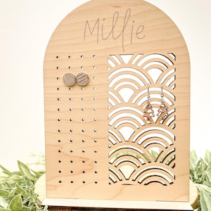 May include: A wooden earring stand with a cutout design of overlapping arcs. The stand has a small shelf at the bottom and a section with holes for stud earrings. The stand is personalized with the name "Millie".