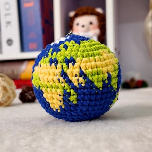 May include: A spherical crochet globe ornament featuring deep blue oceans and textured landmasses in shades of lime green and pale yellow. The handcrafted item includes a small metal loop at the top for hanging, showcasing intricate yarn stitching.