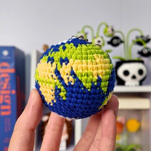 May include: A spherical crochet keychain designed to resemble the planet Earth, featuring intricate stitching in shades of deep blue for the oceans, vibrant lime green for the landmasses, and pale yellow for additional continental details.