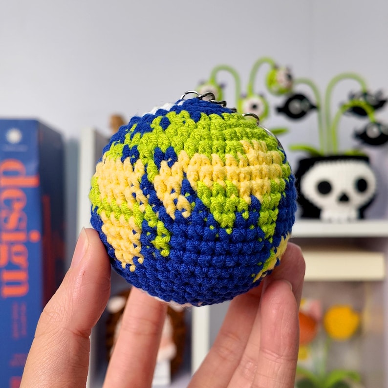 May include: A spherical crochet keychain designed to resemble the planet Earth, featuring intricate stitching in shades of deep blue for the oceans, vibrant lime green for the landmasses, and pale yellow for additional continental details.
