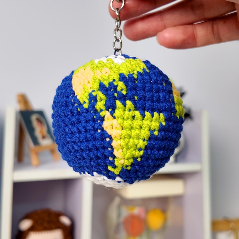 May include: A spherical crochet keychain depicting the Earth, featuring deep blue yarn for the oceans and bright lime green yarn for the continents. The globe is attached to a silver-toned metal chain and a circular split ring for hanging.