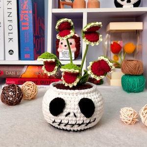 May include: A crochet plant featuring red and green Venus flytrap heads emerging from a white, round pot designed with a black stitched skull face. The plant has multiple stems and textured leaves, sitting as a decorative piece with a stitched mouth.