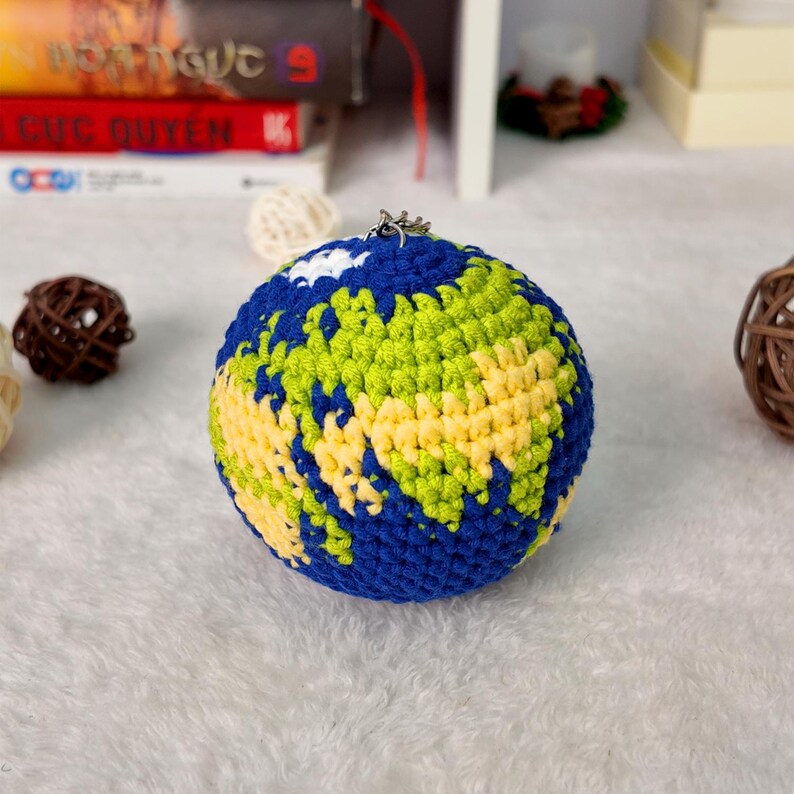 May include: A spherical crochet globe ornament featuring blue yarn for the oceans, light green and yellow yarn for the landmasses, and a small white patch representing a cloud. A silver metal jump ring is attached to the top for hanging.