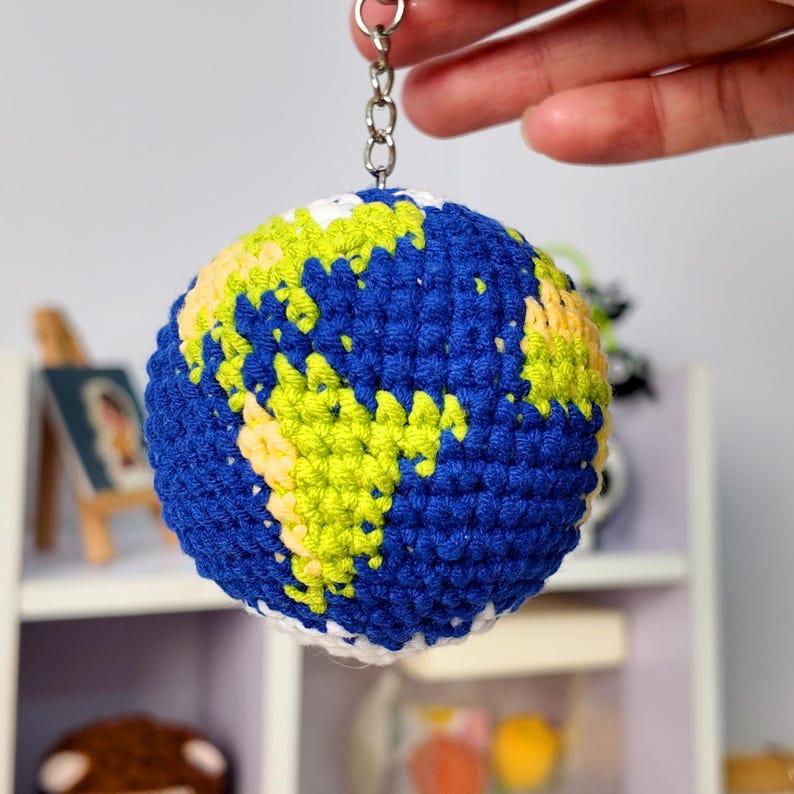 May include: A spherical crochet keychain designed to resemble the planet Earth, featuring deep blue oceans and textured landmasses in shades of lime green and pale yellow, attached to a silver-toned metal chain and a circular split ring.