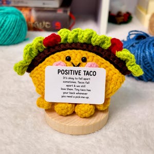 May include: A yellow crocheted taco figure with black eyes, pink cheeks, and green lettuce with red tomato accents. It sits on a round wooden base and holds a white card that reads: POSITIVE TACO. It's okay to fall apart sometimes. Tacos fall apart & we still love them. Tiny taco has your back whenever you need a pick-me-up.