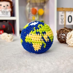 May include: A spherical crochet globe keychain featuring a detailed map design. The surface is crafted with dark blue yarn for the oceans and a combination of light green and pale yellow yarn to represent the continents, topped with a metal ring.