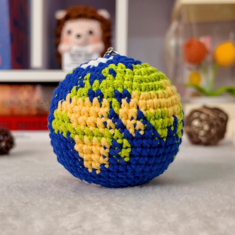 May include: A spherical crochet globe keychain featuring a blue yarn base representing the oceans and yellow and green yarn sections depicting the continents. The item includes a small metal ring at the top for attachment to keys or bags.
