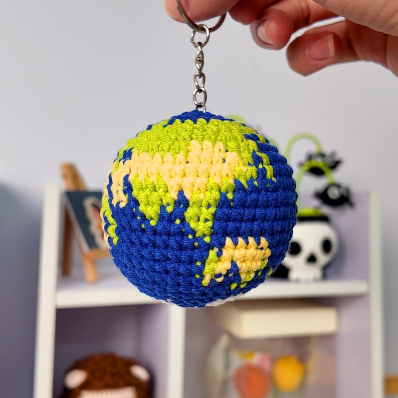May include: A spherical crochet keychain featuring a globe design with deep blue oceans and light green and yellow landmasses. The globe is attached to a silver-toned metal split ring and a short chain, creating a textured, handmade accessory.