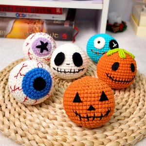 May include: A collection of five spherical crochet toys featuring Halloween-themed designs, including two orange pumpkins with black stitched faces, a white skull with black features, a blue eyeball, and a purple-accented eyeball with red veins.