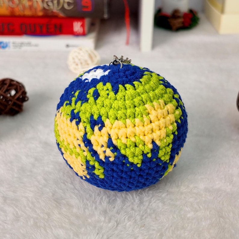 May include: A spherical crochet ornament designed to resemble the Earth, featuring textured blue yarn for the oceans, light green and yellow yarn for the landmasses, and a small white patch for a cloud, topped with a silver metal jump ring.