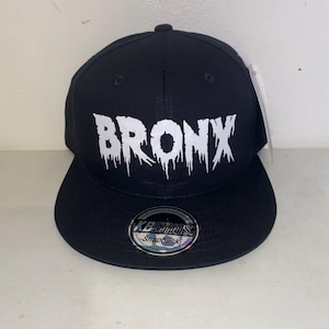 Puede incluir: Gorra snapback negra con la palabra "BRONX" en blanco, con una fuente que gotea en la parte delantera. La gorra tiene una visera plana y una etiqueta. La marca "KBETHOS Snapback" es visible.