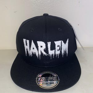 Puede incluir: Gorra snapback negra con la palabra "HARLEM" en blanco, con fuente goteante. La gorra tiene una visera plana y una pegatina "KBETHOS Snapback". Una etiqueta está adjunta al lado.