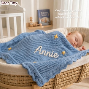 May include: A light blue baby blanket with the name "Annie" embroidered in white. The blanket has a scalloped edge and yellow star accents. The text "Dear Tiny" is in the upper left corner. The blanket is covering a baby in a wicker bassinet.