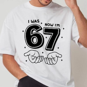 May include: A white t-shirt featuring a black graphic print with the text I WAS NOW I'M 67. The design includes the large number 67, two open hands positioned below the number, and several small four-pointed star accents surrounding the text.