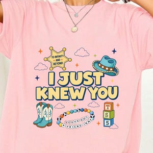 May include: A light pink t-shirt featuring a graphic print with the text I JUST KNEW YOU, a sheriff badge with TO INFINITY AND BEYOND, a blue cowboy hat, cowboy boots, a colorful beaded bracelet with YOU'VE GOT A FRIEND IN ME, and toy blocks.