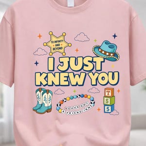 May include: A light pink t-shirt featuring a graphic print with the text I JUST KNEW YOU, a yellow sheriff star with TO INFINITY AND BEYOND, a blue cowboy hat, turquoise cowboy boots, a beaded bracelet with YOU'VE GOT A FRIEND IN ME, and blocks.
